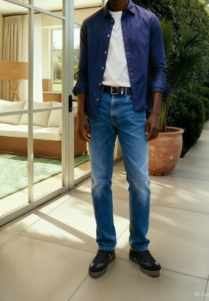 Man wearing blue buttoned shirt over white t-shirt, blue jeans, black belt, and black shoes stands indoors near glass door and potted plant.