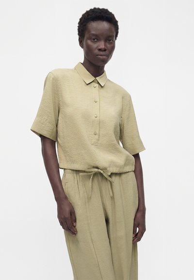 Personne à la peau foncée portant une chemise beige ample à manches courtes avec col et boutons, ainsi qu'un pantalon à cordon assorti, debout devant un fond blanc.