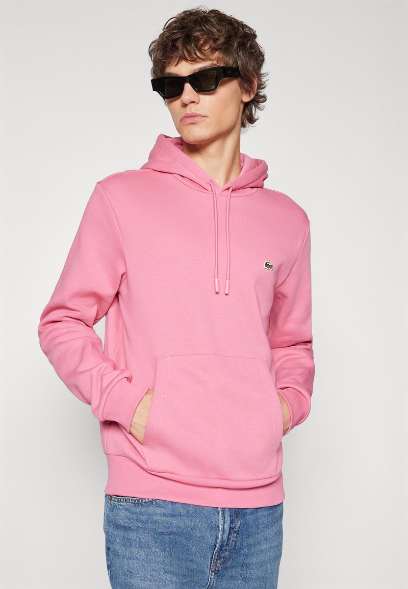 Haut à capuche rose avec poche avant, cordon de serrage, et un petit logo. Porté avec des lunettes de soleil sombres et associé à un jean bleu.