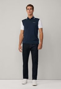 Polo de color azul marino con mangas blancas, de corte ajustado; presenta un cuello tipo polo, logo sutil en el pecho, combinado con jeans oscuros y zapatillas blancas.