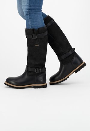 Personne portant des bottes noires mi-mollet avec des sangles à boucle et des semelles couleur fauve, associées à un jean bleu, debout sur un fond blanc.