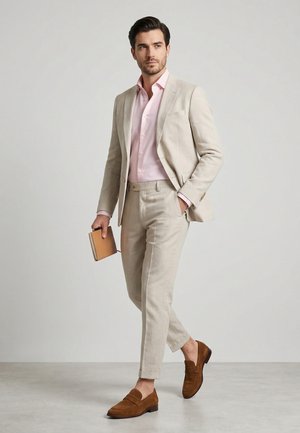 Man walking in beige suit, light pink shirt, brown loafers, holding a tan notebook in right hand against plain background.