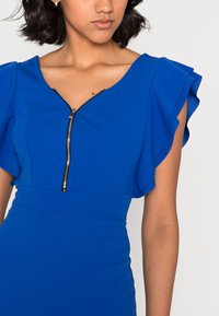 Vestido azul com mangas soltas, apresentando um fecho de correr na frente. Textura suave com uma silhueta ajustada e corpete estruturado.