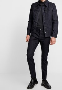 Chaqueta de mezclilla oscura para hombre con frente abotonado, dos bolsillos en el pecho y jeans oscuros a juego. Unos zapatos negros con cordones completan el conjunto.