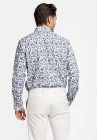Chemise à manches longues en coton blanc avec un motif floral bleu et vert, col boutonné et coupe décontractée, associée à un pantalon blanc.