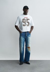Homme portant un T-shirt blanc avec "95" imprimé léopard et "DSQUARED2" en rouge, jean bleu déchiré avec des patchs, casquette noire et chaussures noires, debout immobile.