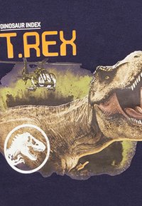Dunkelblauses T-Shirt mit einem großen, detaillierten T. rex-Grafik, einem Hubschrauber darüber, umgeben von grünen Spritzerelementen und einem Dinosaurierlogo.