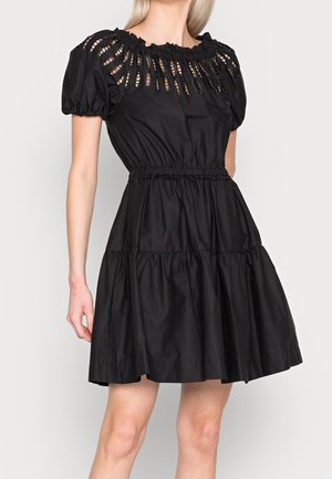 Vrouw draagt een korte zwarte jurk met pofmouwen, een geplooide taille en decoratieve uitsneden langs de halslijn.