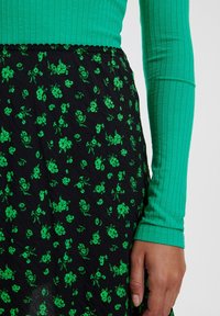 Maglietta verde a maniche lunghe a coste abbinata a una gonna nera con motivo floreale verde vivace. La gonna ha una vita ondulata.