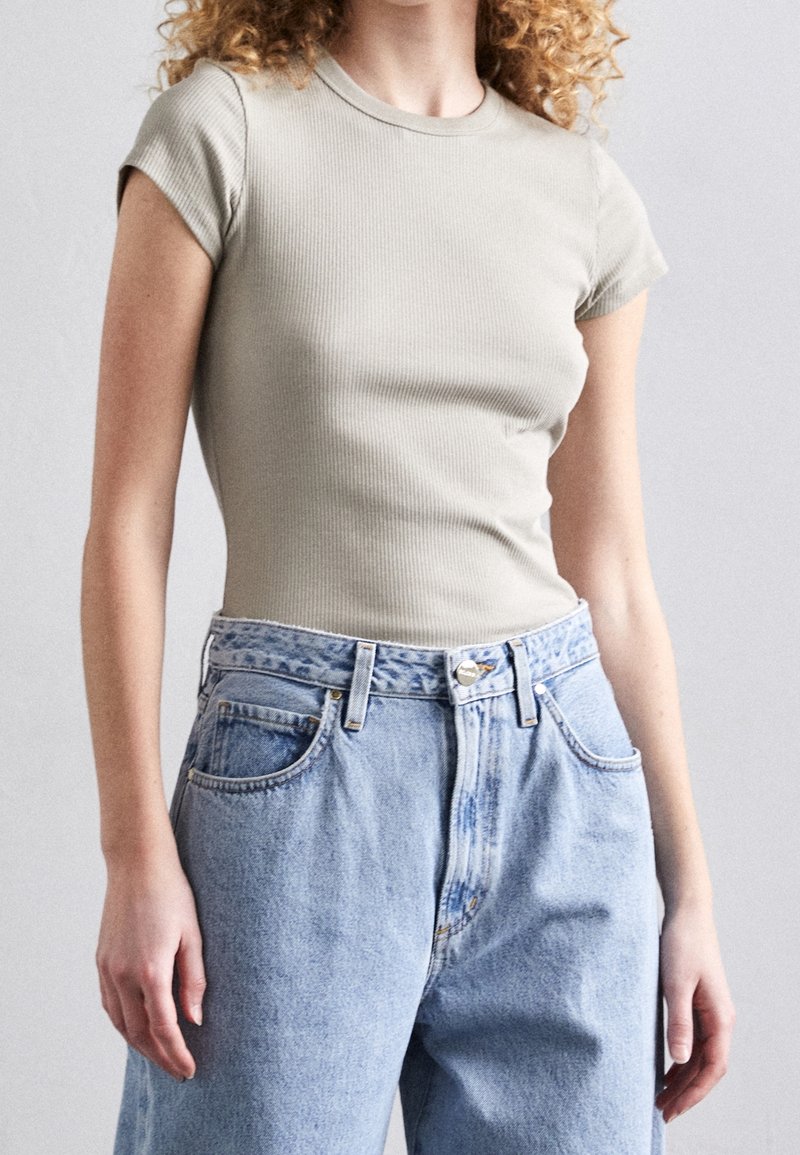 Personne aux cheveux bouclés portant un haut ajusté à manches courtes gris clair et un jean ample taille haute bleu clair sur un fond uni.