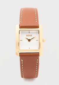 Τετράγωνο χρυσό ρολόι με λευκό καντράν, καφέ δερμάτινο λουράκι με λευκή ραφή και χρυστά δείκτες ώρας και λεπτώ ν που δείχνουν 10:10.