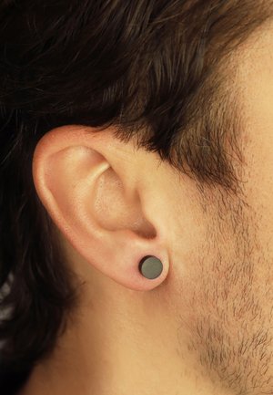 Schwarze, kreisförmige Steckerohrringe aus mattem Material, die in einem durchstochenen Ohr sitzen. Nahaufnahme zeigt Textur- und Formdetails.