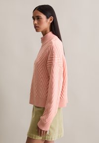 Femme aux longs cheveux bruns portant un pull à col montant texturé rose et une jupe en velours côtelé verte claire, se tenant de profil sur fond uni.