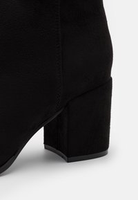 Bottine en daim noir avec talon bloc et couture visible le long de la couture latérale sur fond blanc.