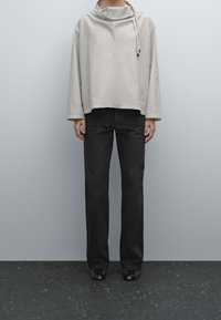 Hellgrauer gestreifter Pullover mit hohem Kragen und Kordelzug, kombiniert mit schwarzen, weit geschnittenen Hosen und glänzenden schwarzen Schuhen.