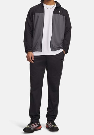 Homme portant une veste zippée noire et grise, un t-shirt blanc, un pantalon noir et des chaussures de sport noires et roses, debout avec une main dans la poche.