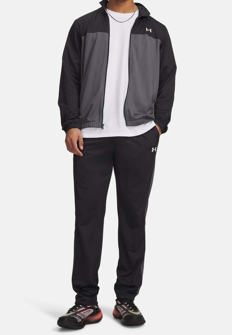 Homme portant une veste zippée noire et grise, un t-shirt blanc, un pantalon noir et des chaussures de sport noires et roses, debout avec une main dans la poche.