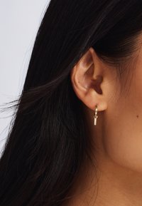 Gouden hoepel oorbel met een kleine verticale staaf hanger. Glad afgewerkt, lichtgewicht ontwerp, en gedragen aan het oor van een model met natuurlijk haar.