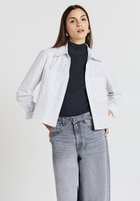 Chemise boutonnée blanche et bleue à rayures avec un col, portée sur un turtleneck côtelé noir et associée à un jean gris taille haute.