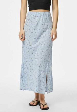 Kvinde iført en lyseblå blomstret maxinederdel med sideslids, sort cropped top og sorte flip-flops, stående mod en ensfarvet baggrund.