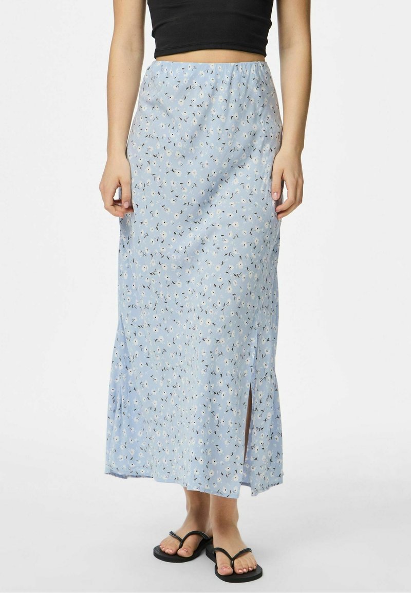 Femme portant une jupe maxi fleurie bleu clair avec une fente latérale, un crop top noir et des tongs noires, debout devant un fond uni.