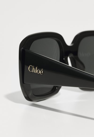 Occhiali da sole neri con montatura quadrata oversize, caratterizzati da una finitura lucida e dal logo dorato "Chloé" sul lato. Lenti scure che offrono protezione dal sole.