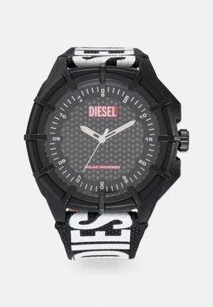 Zwarte zonne-energie aangedreven horloge met een gestructureerde zwarte wijzerplaat, witte markeringen en een zwarte band met gedurfde witte "DIESEL" branding.