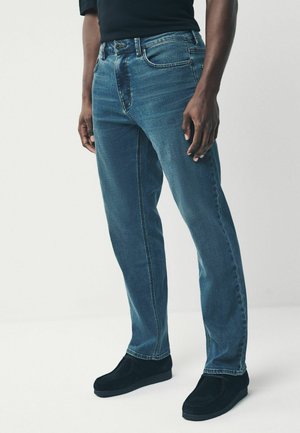 Kék farmernadrág egyenes szárú fazonnal, gombos zárással és elülső zsebekkel. Az anyag enyhe textúrával és kopott hatással rendelkezik.