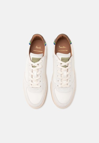 Ζευγάρι λευκά sneakers χαμηλού τύπου με λευκά κορδόνια, ετικέτες γλώσσας ελιάς και καφέ γιακάδες φτέρνας σε ανοιχτό φόντο.
