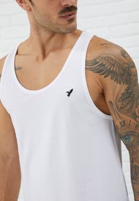 White cotton tank top with a round neckline and black embroidered eagle logo on the left chest. Features a smooth texture and sleeveless design.