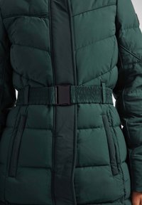 Chaqueta acolchada verde oscuro con un bolsillo lateral, que presenta un cinturón elástico texturizado y un diseño acolchado para el aislamiento. Acabado de tela suave.
