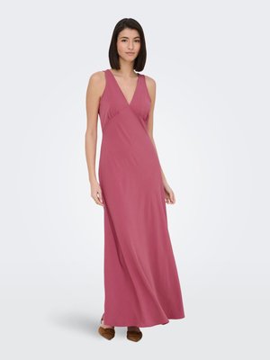 Donna con capelli lisci e scuri che indossa un vestito lungo senza maniche di colore rosa acceso e scarpe basse marroni su sfondo grigio chiaro.