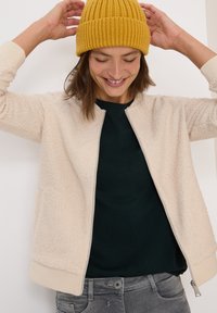 Eine Frau lächelt und richtet ihren senfgelben Strickhut, trägt eine cremefarbene, strukturierte Jacke mit Reißverschluss, ein schwarzes Shirt und graue Jeans vor einem weißen Hintergrund.