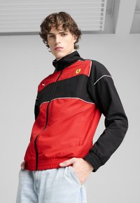 Giacca zip-up rossa e nera con colletto alto, dotata di logo Ferrari, dettagli bianchi e polsini elasticizzati. Materiale leggero e morbido.