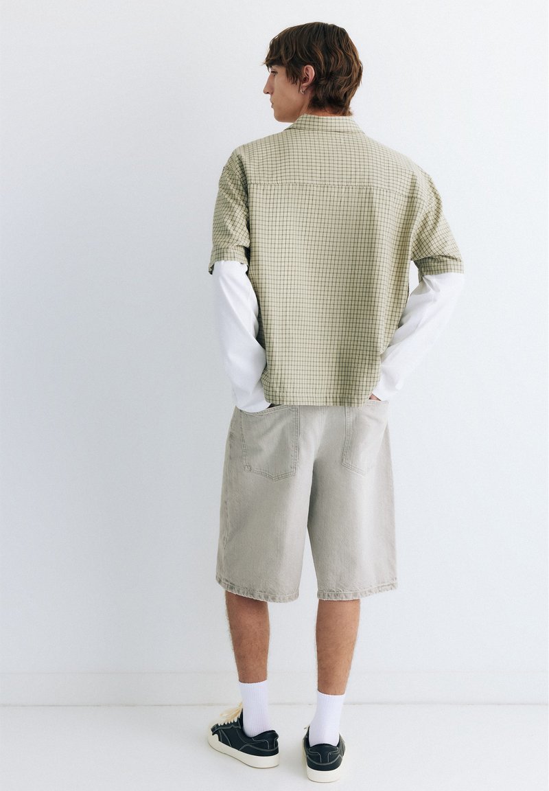 Personne vue de dos portant une chemise légère à manches courtes à carreaux sur un haut blanc à manches longues, un short beige, des chaussettes blanches et des baskets noires.