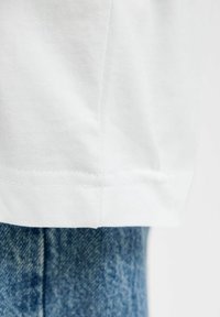 Biely bavlnený lem trička prekrývajúci hornú časť modrých riflí, zobrazujúci textúru látky a detaily šitia.