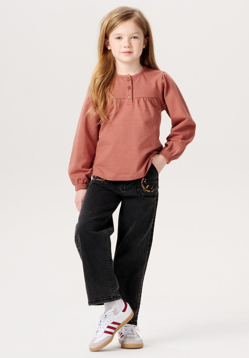 Rostfarbenes karierter Langarmoberteil mit rundem Halsausschnitt und drei Knöpfen. Dunkelblaue Jeans mit floralen Stickereien an den Taschen, kombiniert mit weißen Turnschuhen.