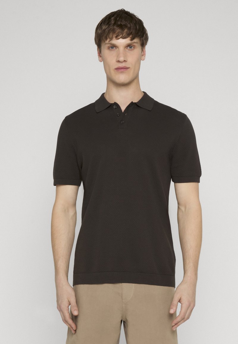 Jeune homme portant un polo noir à manches courtes ajusté et un pantalon beige, debout devant un fond gris clair uni.