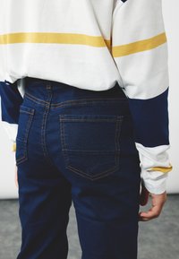 Jeans blu scuro con cuciture arancioni e due tasche posteriori, abbinati a una maglia a maniche lunghe bianca e blu navy con una striscia gialla.