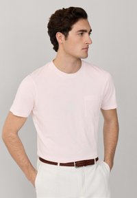 Jeune homme portant un t-shirt rose clair à manches courtes avec une poche poitrine, un pantalon blanc et une ceinture tressée marron, debout les mains dans les poches.