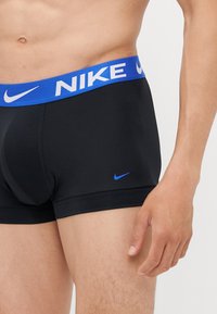 Zwarte boxershorts met een blauwe elastische tailleband waarop in witte tekst "NIKE" staat. Klein blauw logo aan de linkerkant. Glad stoffen textuur.