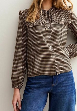 Vrouw met een bruine geruite blouse met een grote gerimpelde kraag en blauwe spijkerbroek, poserend met één hand op haar heup tegen een neutrale achtergrond.