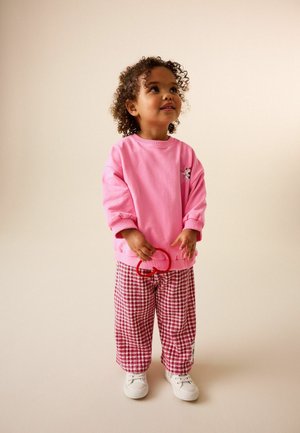 Malé dítě s kudrnatými vlasy, oblečené v růžové mikině a červených kostkovaných kalhotách, držící červený předmět ve tvaru srdce, dívající se vzhůru.