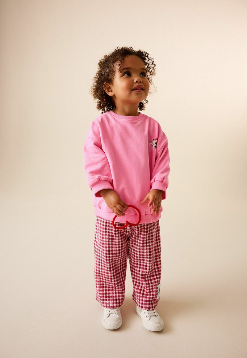 Bambino piccolo con capelli ricci che indossa una felpa rosa e pantaloni a quadri rossi, che tiene in mano un oggetto a forma di cuore rosso, guardando verso l'alto.