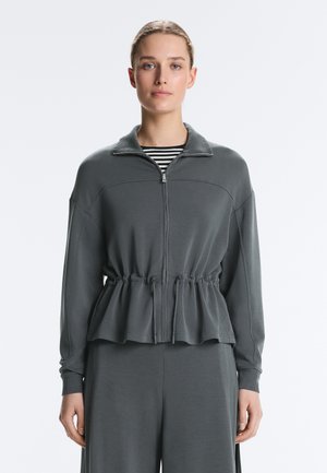 Žena u tamnosivoj jakni na zip s remenom u struku i odgovarajućim hlačama preko crno-bijele prugaste majice, stoji ispred jednobojne pozadine.