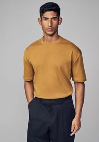 Sennepsgul kortærmet t-shirt med rund hals og afslappet pasform, parret med mørke skræddersyede bukser. Glat stofstruktur.