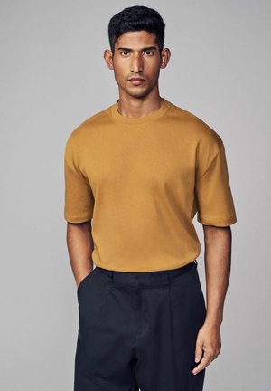 Sennepsgul kortærmet t-shirt med rund hals og afslappet pasform, parret med mørke skræddersyede bukser. Glat stofstruktur.