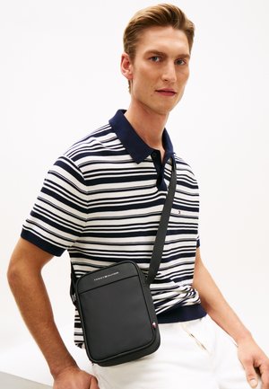 Bărbat purtând pantaloni albi și tricou polo cu dungi bleumarin, având o geantă Tommy Hilfiger neagră purtată peste umăr, pe un fundal alb.