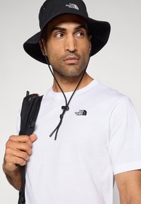 Μαύρο κάπα καπέλο με λογότυπο, λευκό T-shirt με στρογγυλή λαιμόκοψη και μικρό λογότυπο, μαύρα λουράκια σε σακίδιο πλάτης και λεπτομέρεια κορδονιού.