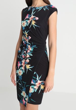 Ærmeløs sort kjole med farverigt blomsterprint, rynket taljedetalje, båret af en stående person med afslappede arme.